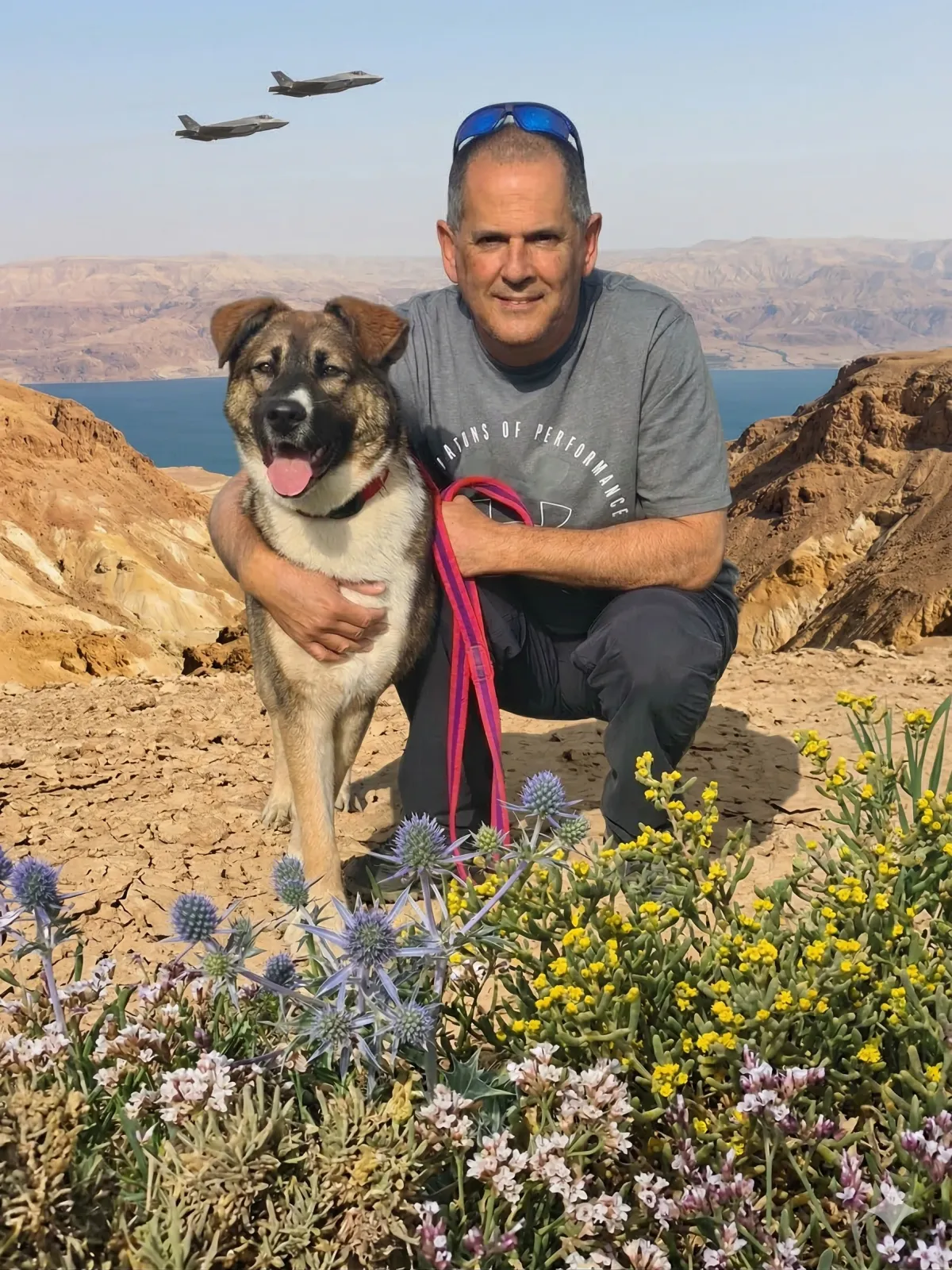 תומר, מאלף כלבים מוסמך, כורע ומחבק כלב רועים צעיר על רקע נוף מדברי